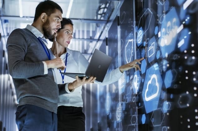 A man and a woman in a server room holding a lap top, pointing at the servers