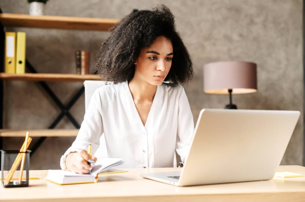 Young woman working in her office at a desk on her lap top and writing notes in her notepad