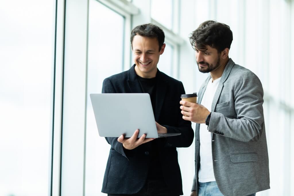 Two male professional standing in hallway at office holding coffee cups and looking at a lap top